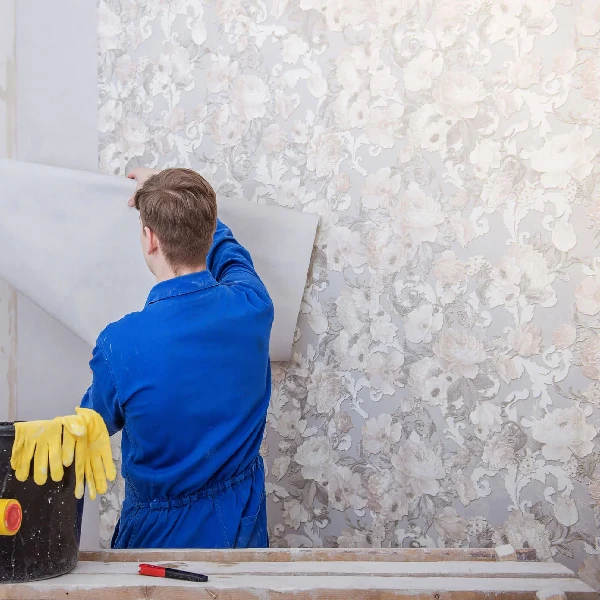 blue jumpsuit applies a floral-patterned wallpaper sheet to a wall.