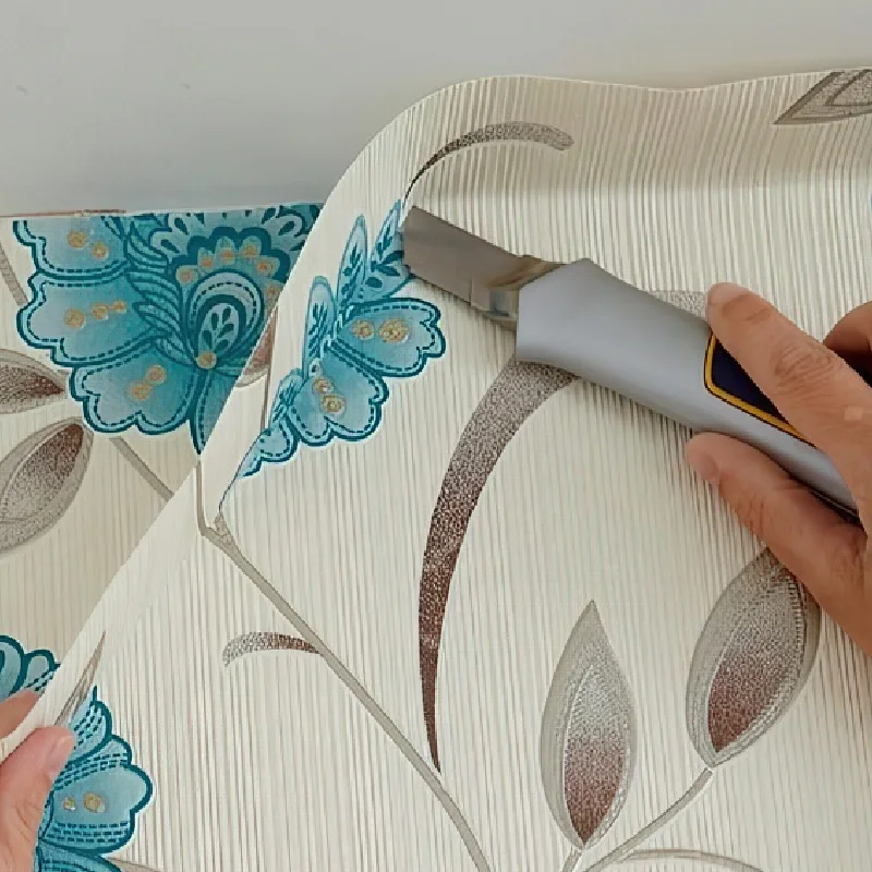 person using a utility knife to trim a floral-patterned wallpaper sheet.